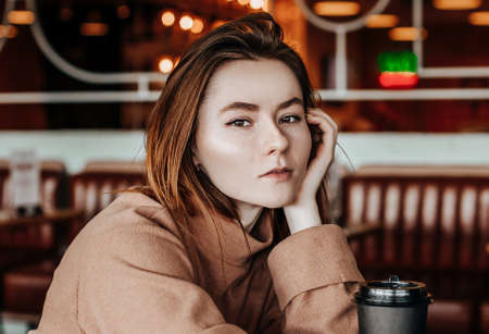 Stylish girl sits in a cafe at the table and drinks coffee. Coffee to go in a cardboard cup. Woman with red hair in a beige warm suit in a cozy atmosphere. Modern interior. Calm and pleasant pastime.の写真素材