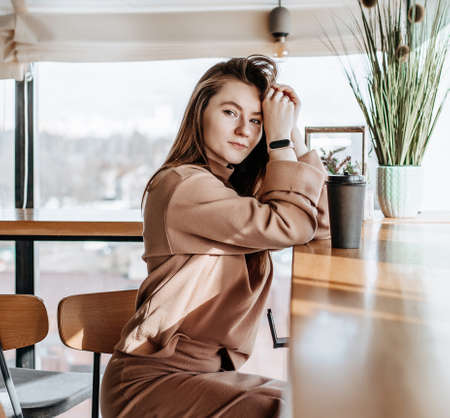 Stylish girl sits in a cafe and drinks coffee. Coffee to go in a cardboard cup. Woman with ginger hair in a beige warm suit in a cozy atmosphere. Modern interior. Calm and pleasant pastimeの写真素材