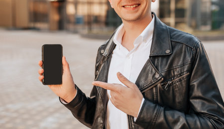 Handsome with a smartphone on the street of a big city. Businessman points finger at phone screen on urban backgroundの写真素材