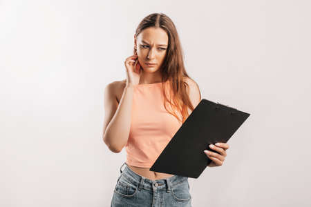 Girl student frowns with displeasure and reads information in documents. Beautiful young business woman holds a clipboard with mockup on gray background. Study and business concept. Online learningの写真素材