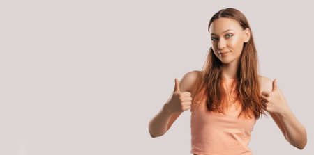 Beautiful young girl smiling and shows thumbs up gesture with two hands on a white isolated background. Positive woman points to an idea, a place for advertisingの写真素材