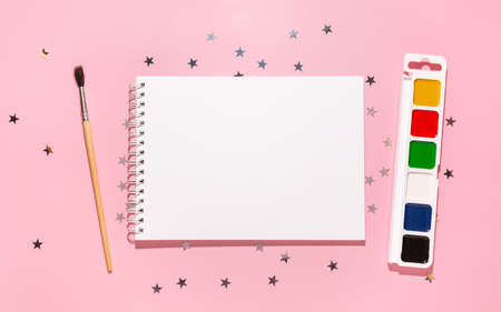 Empty notebook with paints, brush and confetti stars pours on a pastel pink background, top view. Flat lay. Holiday, greeting or good news concept. Mockup template. top view. Blank notepadの写真素材