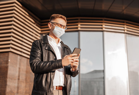 Handsome man in a protective mask on his face with glasses with a smartphone on the street of a big city. Businessman talking on the phone on urban backgroundの写真素材