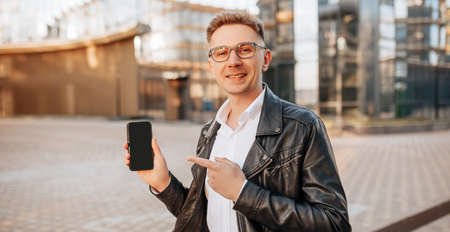 Handsome man in glasses with a smartphone on the street of a big city. Businessman points finger at phone screen on urban backgroundの写真素材