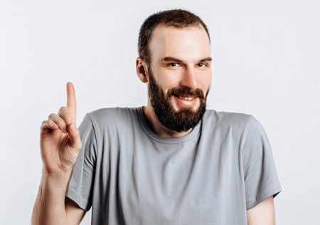 Portrait of cheerful young handsome man smiling looking at camera pointing finger upwards on white background with space for advertising mock upの写真素材