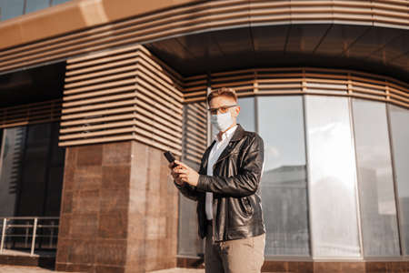 Handsome man in a protective mask on his face with glasses with a smartphone on the street of a big city. Businessman talking on the phone on urban backgroundの写真素材
