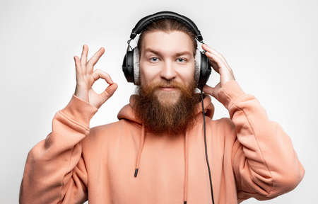Scandinavian handsome man shows ok gesture smiles and listens to music in professional headphones isolated on gray background. Happy guy with ginger hairstyle and beard. Modern digital technologiesの写真素材