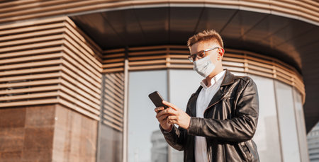 Handsome man in a protective mask on his face with glasses with a smartphone on the street of a big city. Businessman talking on the phone on urban backgroundの写真素材