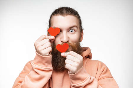 Portrait of happy handsome man with ginger hairstyle and beard on gray background. Attractive scandinavian man being playful and holding red hearts over eyesの写真素材