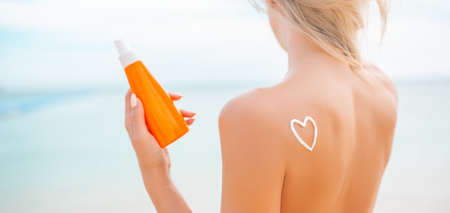 Woman applying sun cream on tanned body In form heart love to protect her skin. Girl using sunscreen. Female holding suntan lotion, moisturizing sunblock on sunny day with blurry sea in the backgroundの写真素材