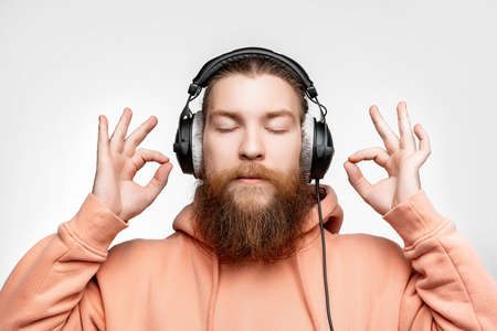 Scandinavian handsome man shows ok gesture keep calm and listens to music in professional headphones isolated on gray background. Happy guy with ginger hairstyle and beard. Modern digital technologiesの写真素材