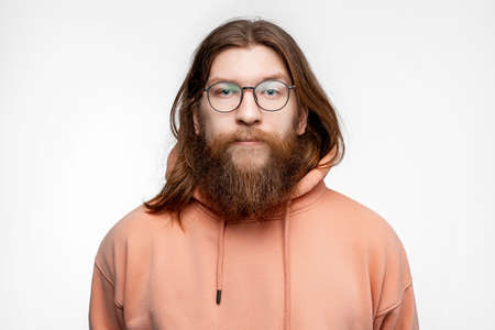 Scandinavian handsome serious man with glasses and face hair isolated on gray background in orange sweatshirt. Calm nordic guy with long ginger hair and beardの写真素材