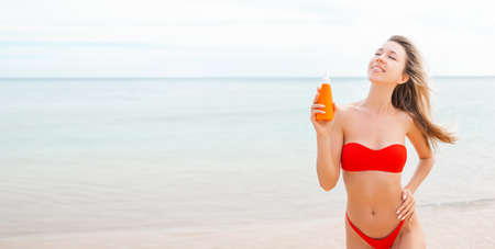 Woman applying sun cream on tanned body In form heart love to protect her skin. Girl using sunscreen. Female holding suntan lotion, moisturizing sunblock on sunny day with blurry sea in the backgroundの写真素材