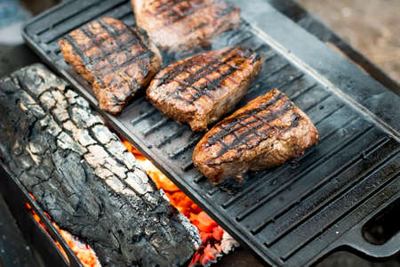 Beef steaks grilling on a cast iron plate on a camp fire. Campfire cooking. Outdoor BBQ. Selective focusの写真素材