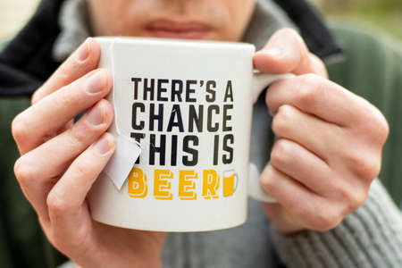 Autumn winter camping life concept. A man in a grey sweater and green vest drinking tea for the white cup with funny phrase on itの写真素材