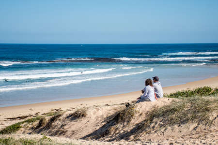 Bendalong, Australia 2020-06-06 An adult couple looking over the ocean while sitting on sand dunes on a sunny day. Bendalong Beach, NSW, Australiaのeditorial素材