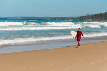 Bendalong, Australia 2020-06-06 Adult man surfer in red wetsuit carrying surfboard on a sandy beachのeditorial素材