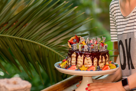 Selective focus image of hands holding layered cake decorated with sparkles, happy birthday candles, chocolate glaze, strawberries, blueberries, carambola star fruits on tropical backgroundの写真素材