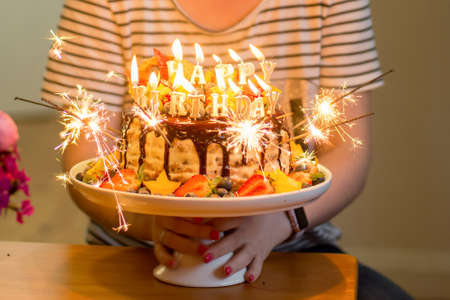 Disfocused image of hands holding delicious handmade layered cake decorated with sparkles, happy birthday candles, chocolate glaze, strawberries, blueberries, carambola star fruit and ice cream coneの写真素材