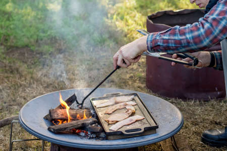 Breakfast camp cooking. Man grilling crispy bacon on a cast iron plate over the camp fireの写真素材