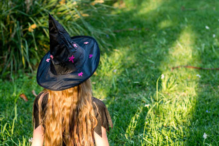 Causcasian blond girl with long hair in halloween witch costume sitting on a green grass