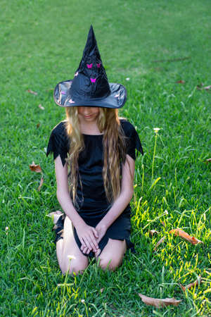 Causcasian blond girl with long hair in halloween witch costume sitting on a green grassの写真素材