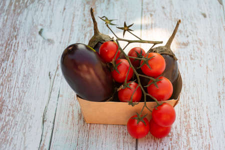 Eggplant aubergines and red ripe cherry tomatoes in a paper box on a wooden table in the gardenの写真素材
