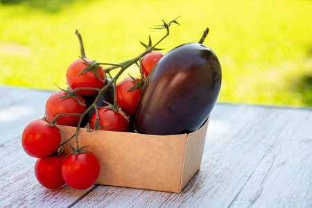Eggplant aubergines and red ripe cherry tomatoes in a paper box on a wooden table in the gardenの写真素材