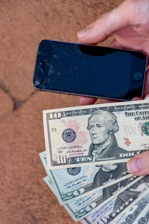 Sydney, Australia - 2019-11-01 Hand holding a phone with badly smashed screen and US dollars to pay...