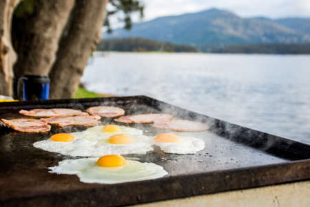 Breakfast camp cooking, Grilling eggs and bacon on the bbq plate with beautiful nature landscape on background.の写真素材