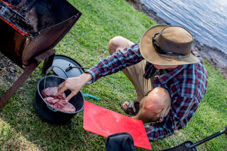 Dutch oven campfire cooking process. A man put lamb shoulders in a cast iron camp oven. Camping life.の写真素材