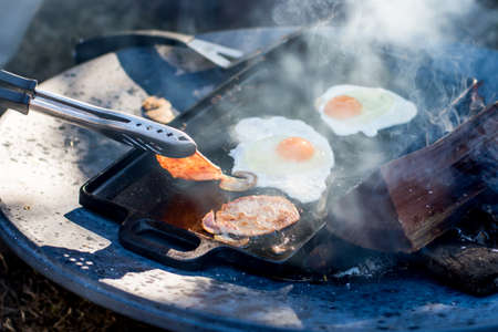 Breakfast camp cooking. Grilling crispy bacon and eggs on a cast iron plate over the camp fire.の写真素材