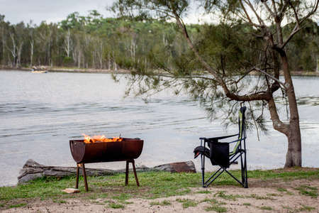 Campfire in a half barrel fire pit and empty campchair on the lake. Camping life.の写真素材