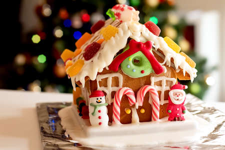 Homemade gingerbread house decorated with icing, sweets , figurines, lollies and candy canes with blurred Christmas tree on background.の写真素材