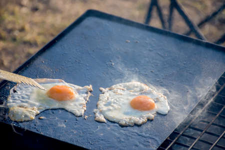 Disfocused shot of breakfast camp cooking. Grilling crispy bacon and eggs on a cast iron plate over the camp fireの写真素材