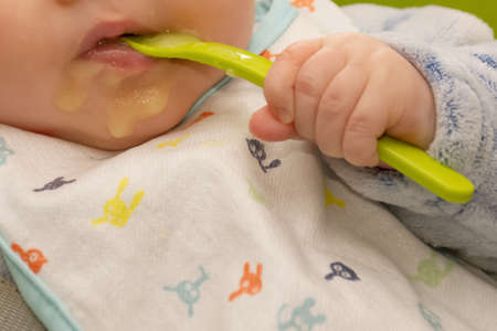 Baby holds a spoon and put it in his mouth with his little hand. Baby first soilds. Weaning.の写真素材
