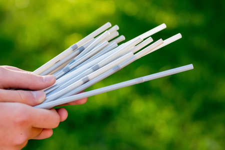 Hand holding white plastic straws on green grass background. Plastic pollution, environmental waste. Say no to plasticの写真素材