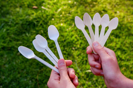 Hands holding disposable wooden teaspoon and plastic teaspoons on a green grass background. Reduce Reuse Recycle. Say no to plastic cutleryの写真素材