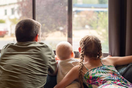 A blond girl, her baby brother and their father looking through the window. Family coronavirus sellf isolation lockdown conceptの写真素材
