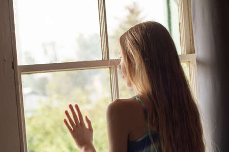 Lonely teenage girl waiting near the hazy window inside the room. Self isolation lockdown...