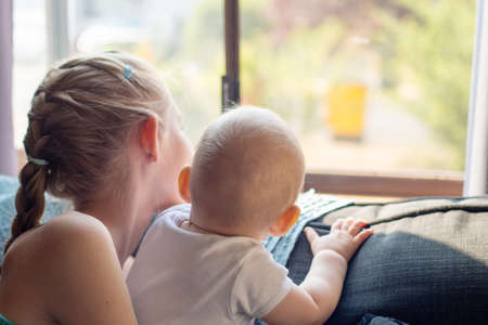 A blond girl holds her baby brother near the window. Self isolation lockdown coronavirus concept.の写真素材