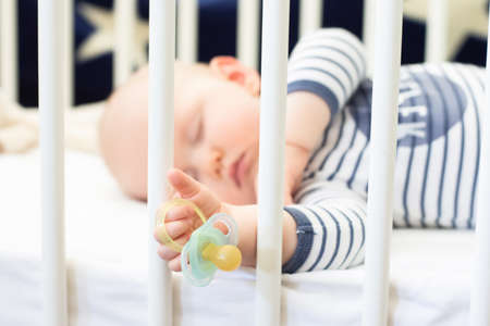 Little baby boy peacefully sleeping in the bed holding pacifier dummy in his hand. Selective focus.の写真素材
