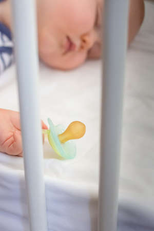 Little baby boy peacefully sleeping in the bed holding pacifier dummy in his hand. Selective focus.の写真素材