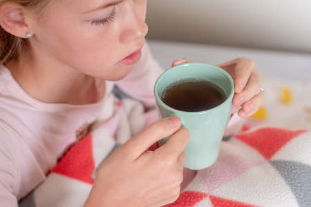 Sick teenage girl sitting on the bed covered with blanket drinking hot tea. Healthcare, Herbal medicineの写真素材