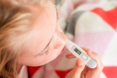 Defocused image of sick teenage girl sitting on the bed covered with blanket measure body temperature. Fever, cold and COVID-19 conceptの写真素材