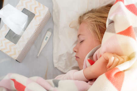 Sick teenage girl lying on the bed covered with blanket with thermometer. Covid-19 symptomsの写真素材