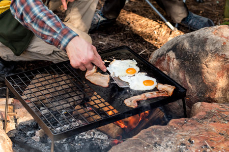 Breakfast camp cooking. Grilling crispy bacon and eggs on a cast iron plate over the camp fire. Camping lifesyleの写真素材