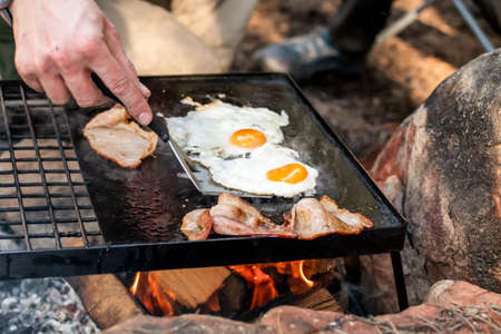 Breakfast camp cooking. Grilling crispy bacon and eggs on a cast iron plate over the camp fire. Camping lifesyleの写真素材