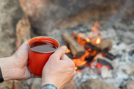 Camping lifestyle concept. Hands holding red enamel mug near a campfire. Hikingの写真素材