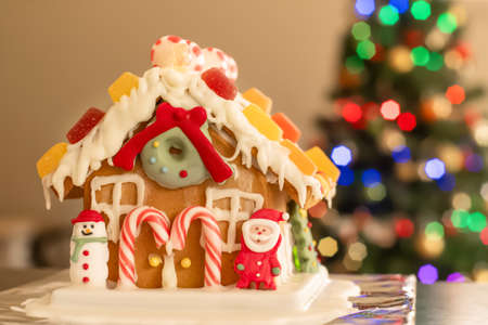 Homemade gingerbread house decorated with icing, sweets , figurines, lollies and candy canes with blurred Christmas tree on background.の写真素材